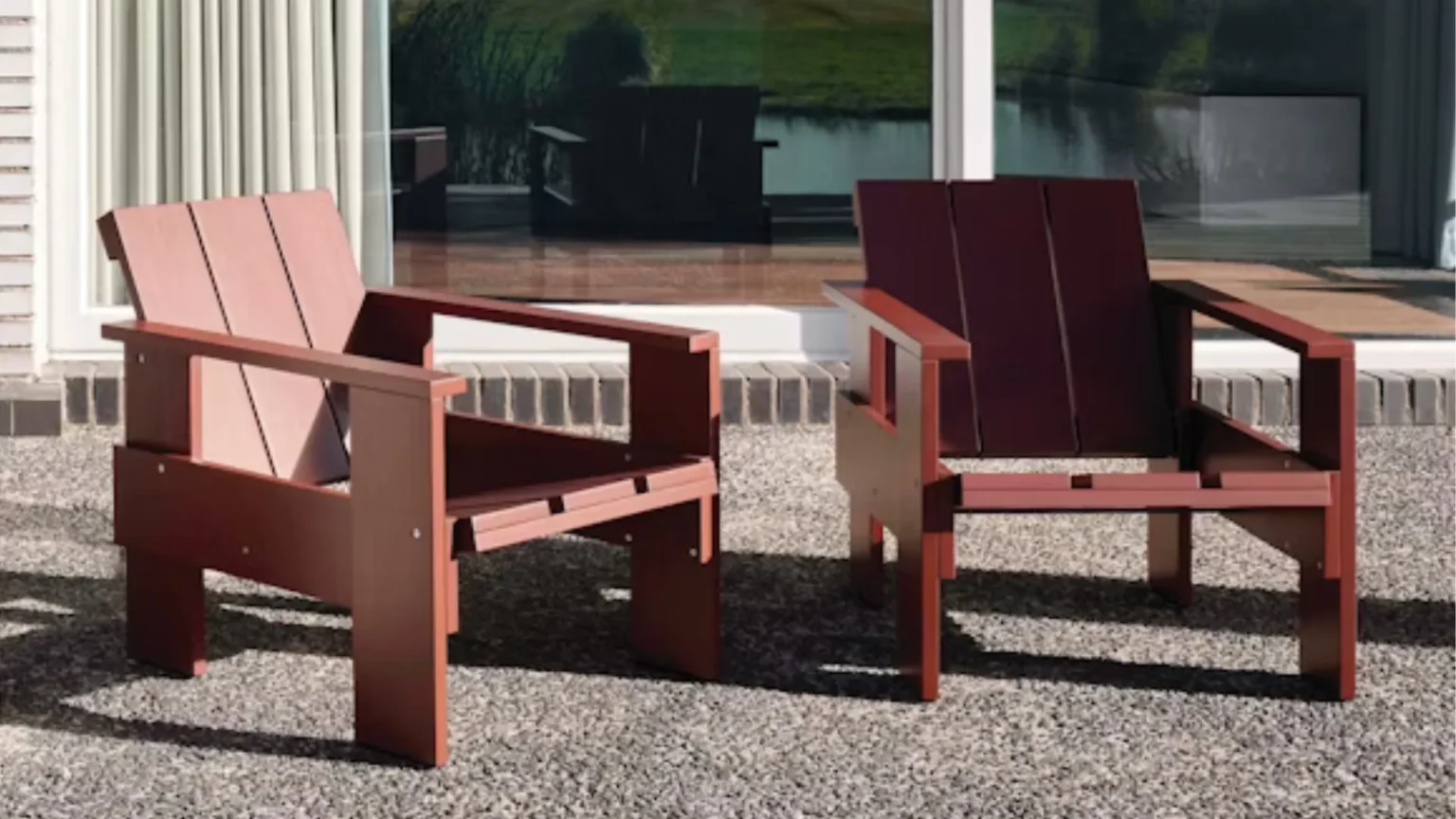 Two wooden lounge chairs with a modern design placed on a gravel patio in front of a glass wall