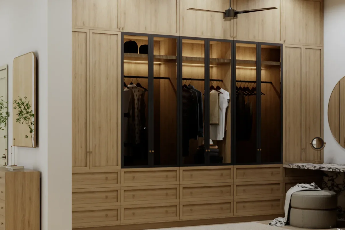 Light oak floor-to-ceiling wardrobe wall with black-framed glass doors showing LED-lit hanging clothes and drawers, marble vanity desk and round mirror in corner