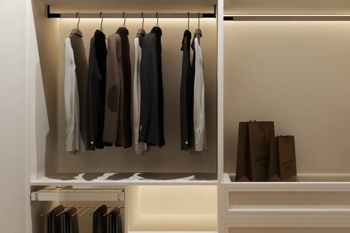 White built-in wardrobe interior with warm LED strip lighting along the top rail, neutral and dark hanging garments, a pull-out trouser rack below, and open shelf niche with brown shopping bags