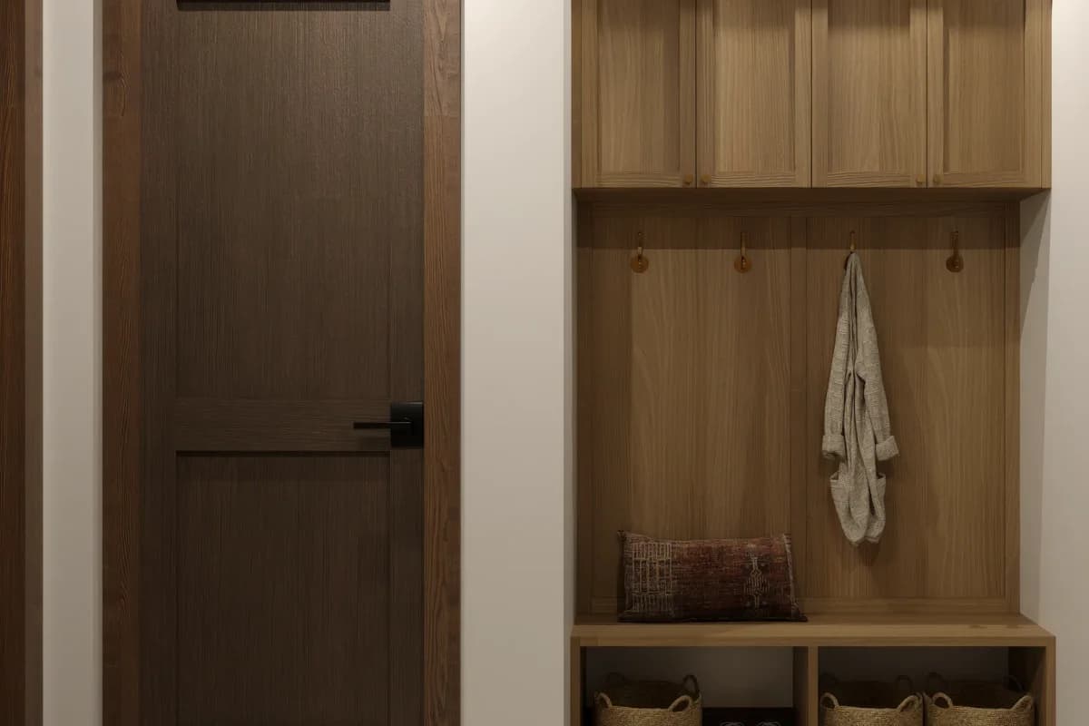 Natural oak mudroom built-in with upper closed cabinets, brass round hooks, open bench cubby, and woven storage baskets below