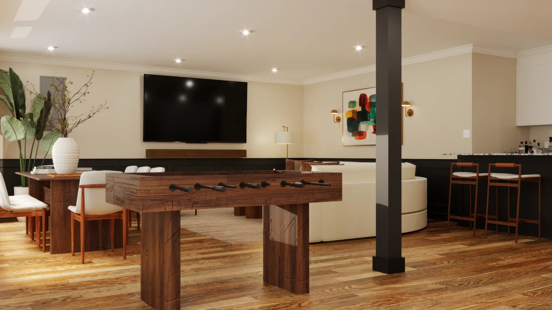 Basement game room with walnut foosball table centered on hardwood floor, cream curved sectional, wall-mounted TV above walnut media console, abstract multicolor painting with brass sconces, black column, and bar counter with wood stools.