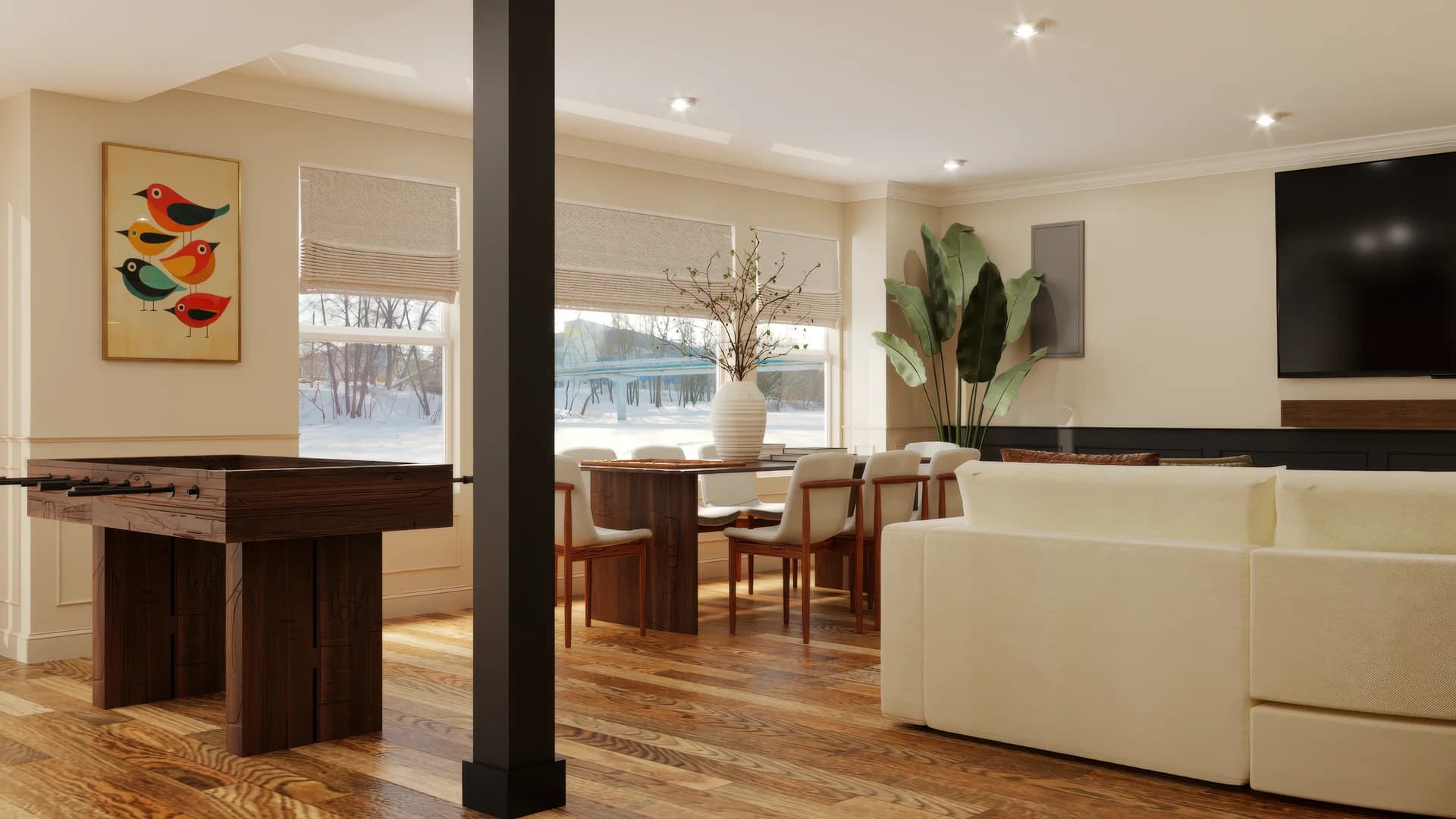 Basement open-plan space with walnut foosball table, black structural column, cream sectional sofa, wall-mounted TV, walnut dining table with tan chairs, large plant, white vase with branches, and colorful bird artwork on cream walls.