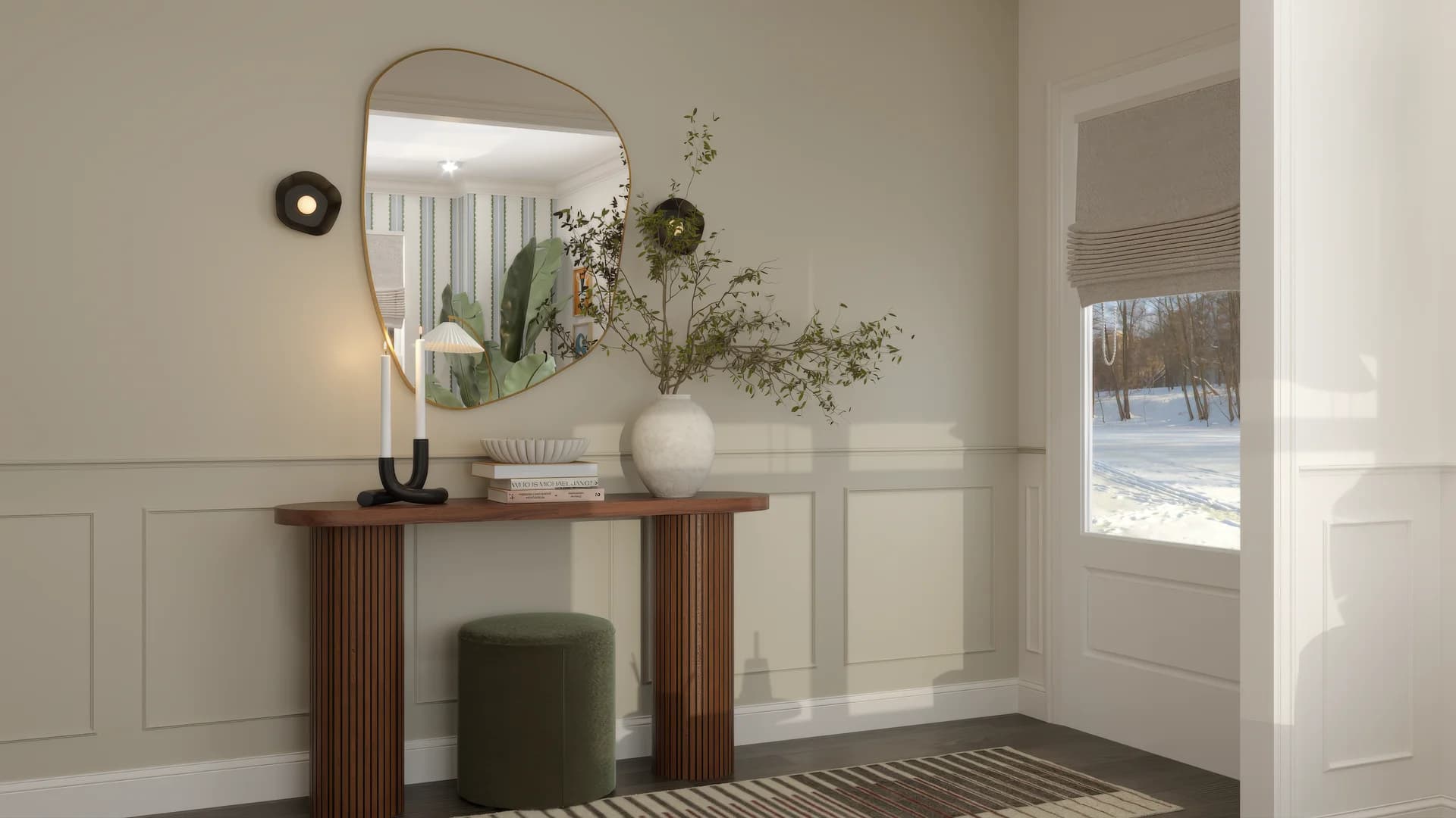 Entryway with organic-shaped gold brass mirror, reeded walnut console table topped with white ceramic vase, branching greenery stems, stacked books, shell bowl, black taper candle holder, olive green boucle ottoman beneath, and wainscoting walls.