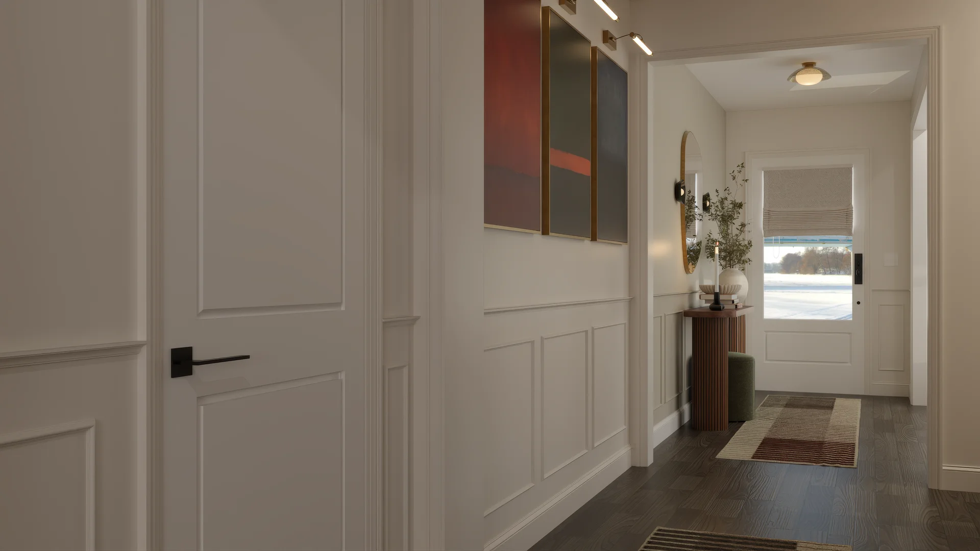 Hallway with white wainscoting panels, abstract three-panel artwork in burgundy and slate tones with brass picture lights, dark hardwood floors, geometric striped runner rug, round gold mirror, fluted wood pedestal table, and white front door.