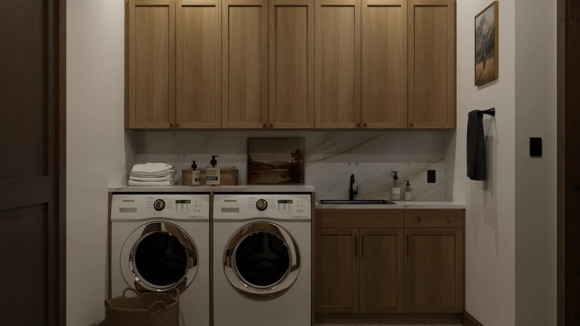 Farmhouse laundry room with front-load washer and dryer, warm oak cabinets, calacatta marble backsplash, utility sink, woven hamper basket, and landscape painting