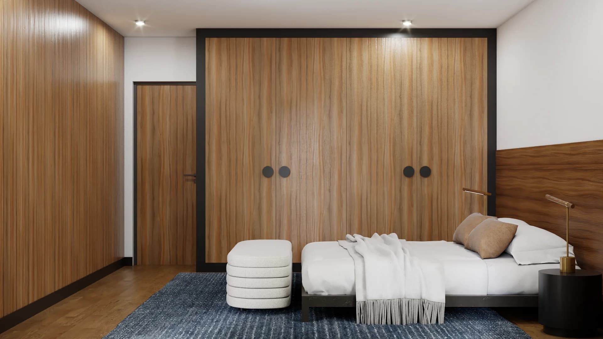 Walnut wardrobe wall with black handles flanking a platform bed, cream ottoman, brass reading lamp, and navy rug on parquet floor