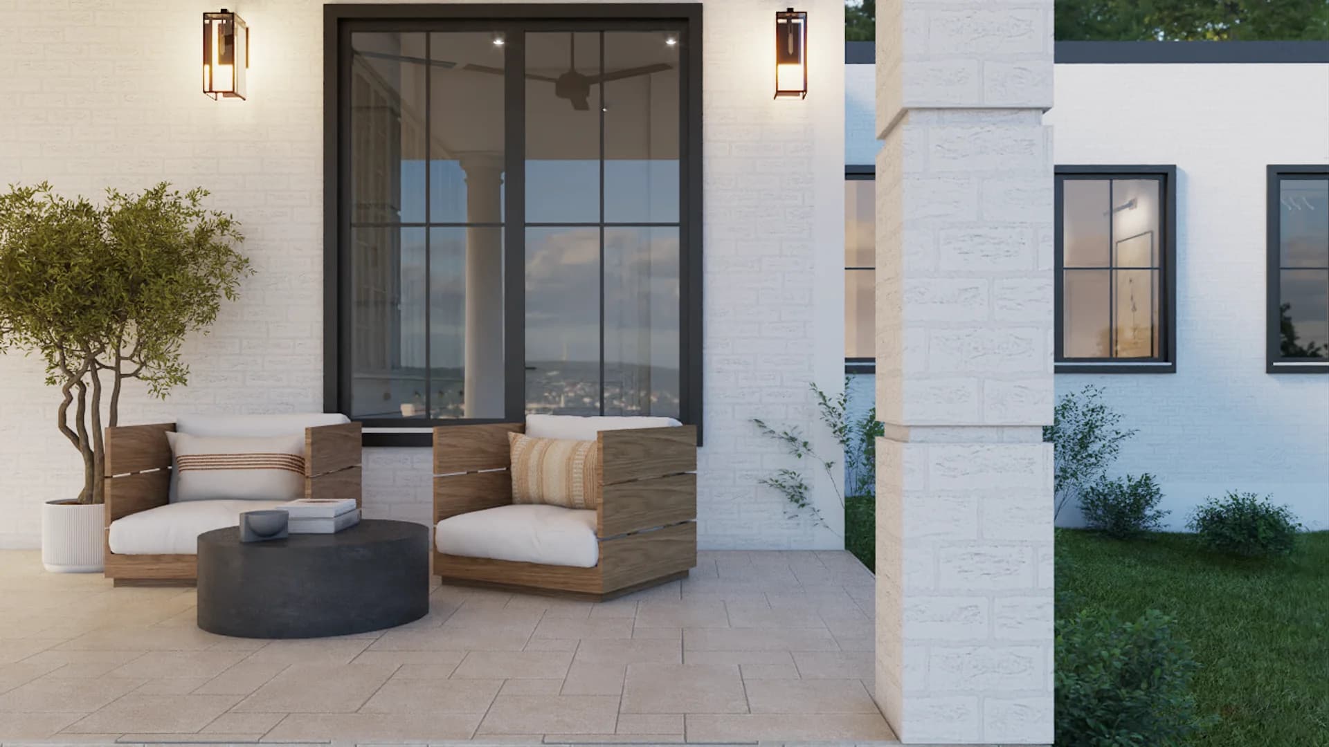 Covered outdoor patio with teak lounge chairs and cream cushions, round dark concrete coffee table, white brick wall, black iron sconces, and potted olive tree at dusk by Debora Interiors