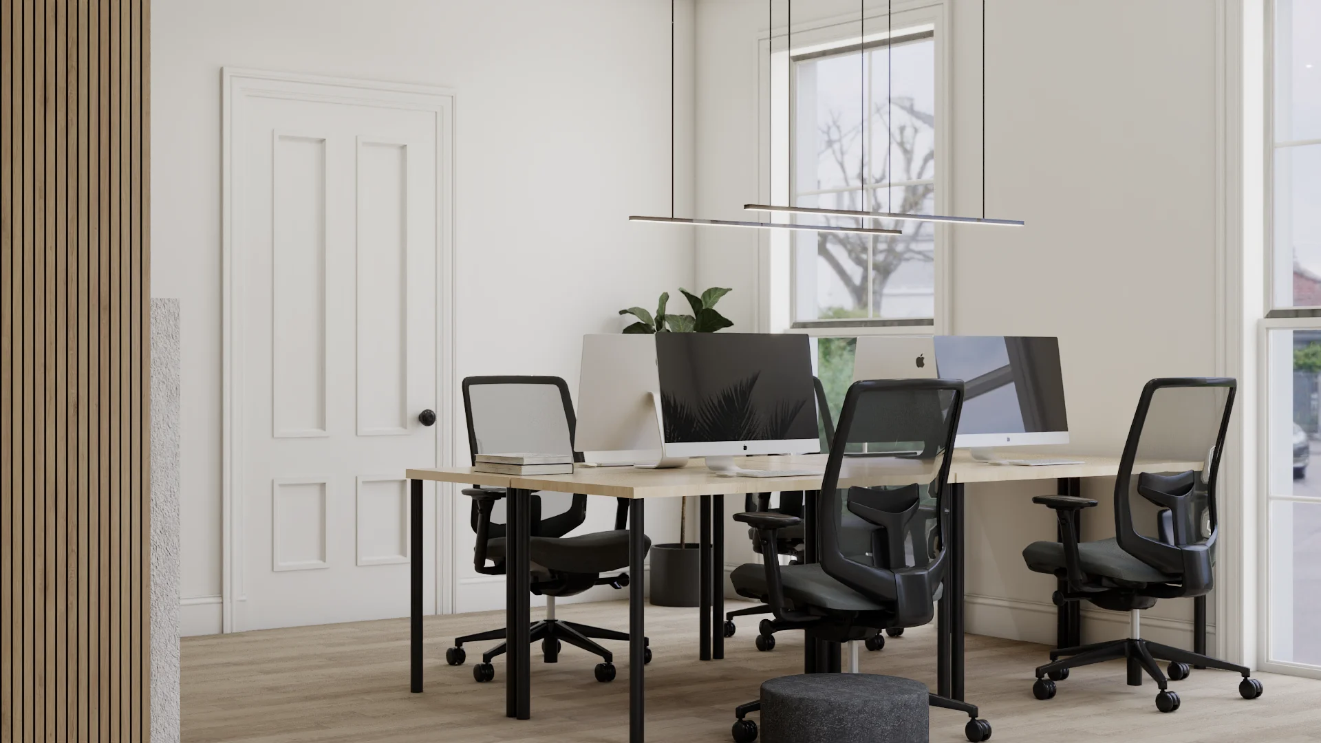 Four-desk shared bench workspace with maple tops and black metal legs, black ergonomic task chairs, iMac monitors and laptop, double linear pendant lights, vertical oak slat wall panel at left, white paneled door, tall windows with street view