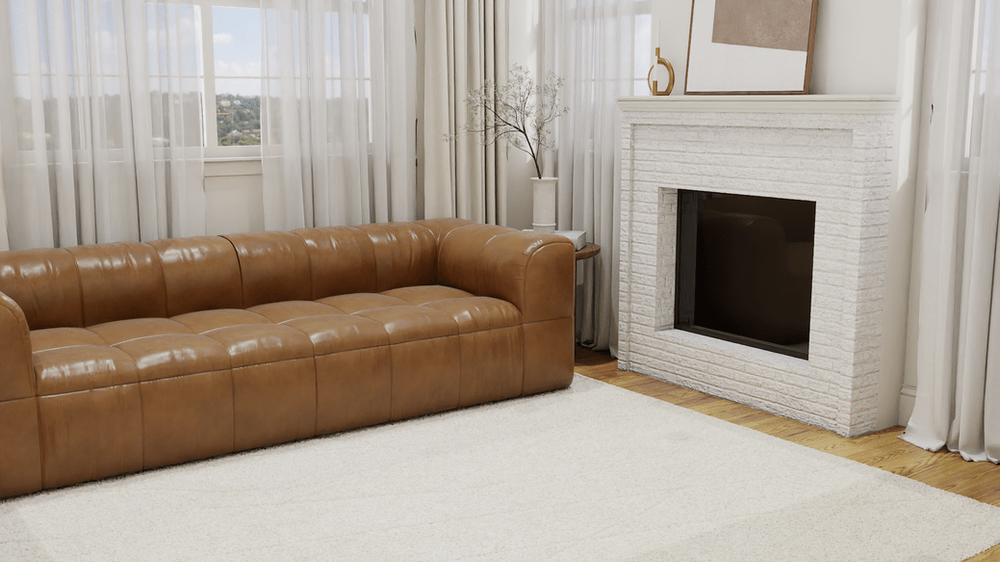 A cozy living room featuring a modern, tan leather tufted sofa placed on The Diamond Drift Shag Rug by West Elm, which adds texture and warmth with its thick, plush pile and diamond pattern in a light beige hue. The sofa is positioned in front of a white brick fireplace with a minimalist mantel displaying a small vase with delicate branches and a gold decorative object. The room is softly illuminated by natural light streaming through large, sheer curtains that frame a view of a distant landscape outside the window, designed by Debora