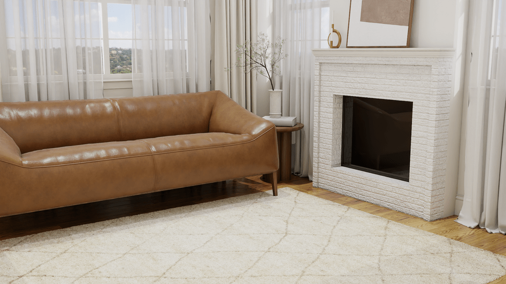A stylish living room showcasing a sleek, tan leather sofa resting on The Afella Rug by Lulu and Georgia. The rug features a delicate diamond pattern and a luxurious, cream-colored pile that complements the room's aesthetic. The sofa faces a white, textured brick fireplace adorned with minimal decor, including a small vase with twigs and a gold ornament. Natural light filters through sheer curtains, creating a serene ambiance and offering a glimpse of the outdoor scenery