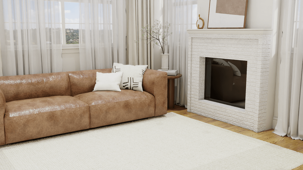The room includes a white, textured brick fireplace with minimalist decor, including a small vase with delicate branches and a gold ornament. Large, sheer curtains allow natural light to gently illuminate the space, adding to the serene and airy ambiance while offering a view of the outside landscape, tan leather sofa situated on The Chunky Wool - Jute Rug by Pottery Barn.