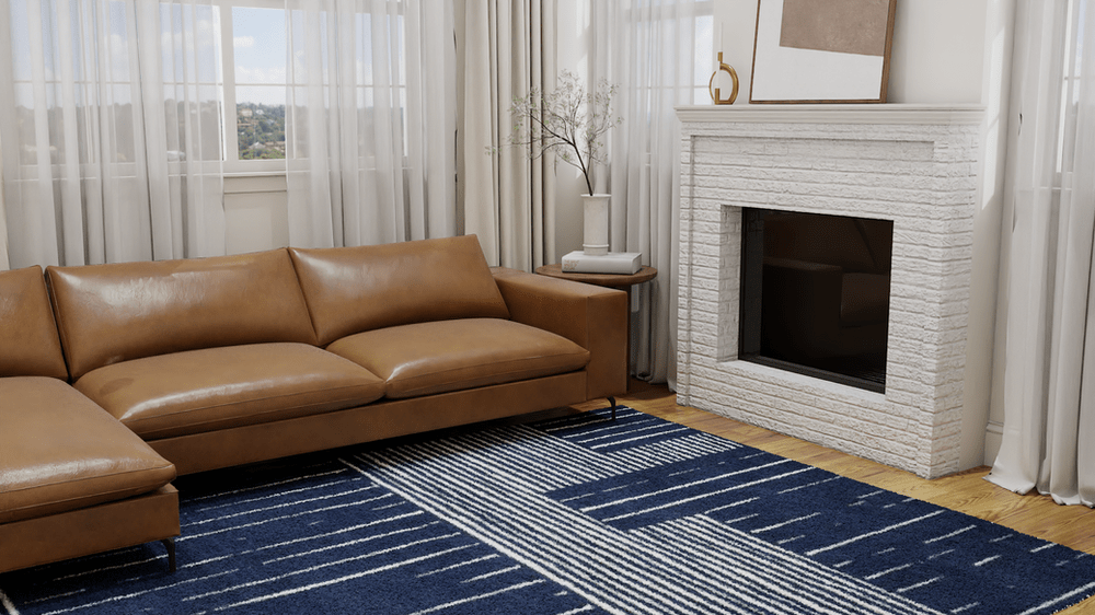 A modern living room showcasing a sleek, tan leather sectional sofa arranged on The Painted Mixed Stripes Rug. This rug features a bold design with a dynamic mix of navy and white stripes, adding a striking contrast and contemporary flair to the space. The room is complemented by a white brick fireplace with minimalist decor, including a vase of branches and a gold ornament. Sheer curtains allow natural light to bathe the room, creating an airy and bright environment with views of the landscape outside