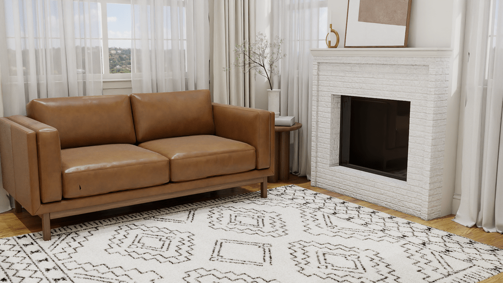 A cozy living room featuring a modern, tan leather loveseat set against a backdrop of sheer curtains that let in ample natural light. The floor is anchored by The Gray Diamond Geometric Area Rug, which adds a touch of sophistication with its intricate, gray diamond patterns on a white background. The space is enhanced by a white brick fireplace adorned with simple decor, including a vase with twigs and a gold ornament, contributing to a clean, minimalistic aesthetic