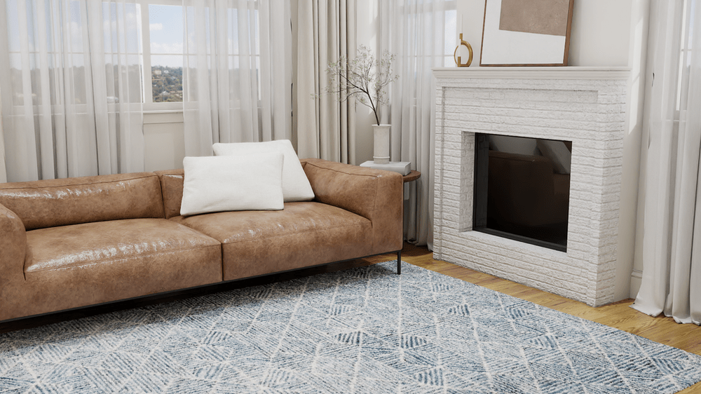 A contemporary living room featuring a stylish, tan leather sofa adorned with white throw pillows. The sofa rests on The Abstract ABT763M Blue Rug by Safavieh, which boasts a subtle, modern pattern in shades of blue and gray, adding depth and texture to the space. The room also includes a white brick fireplace with minimalist decor, such as a vase of branches and a gold ornament. Sheer curtains allow natural light to flood the room, enhancing its bright and airy feel